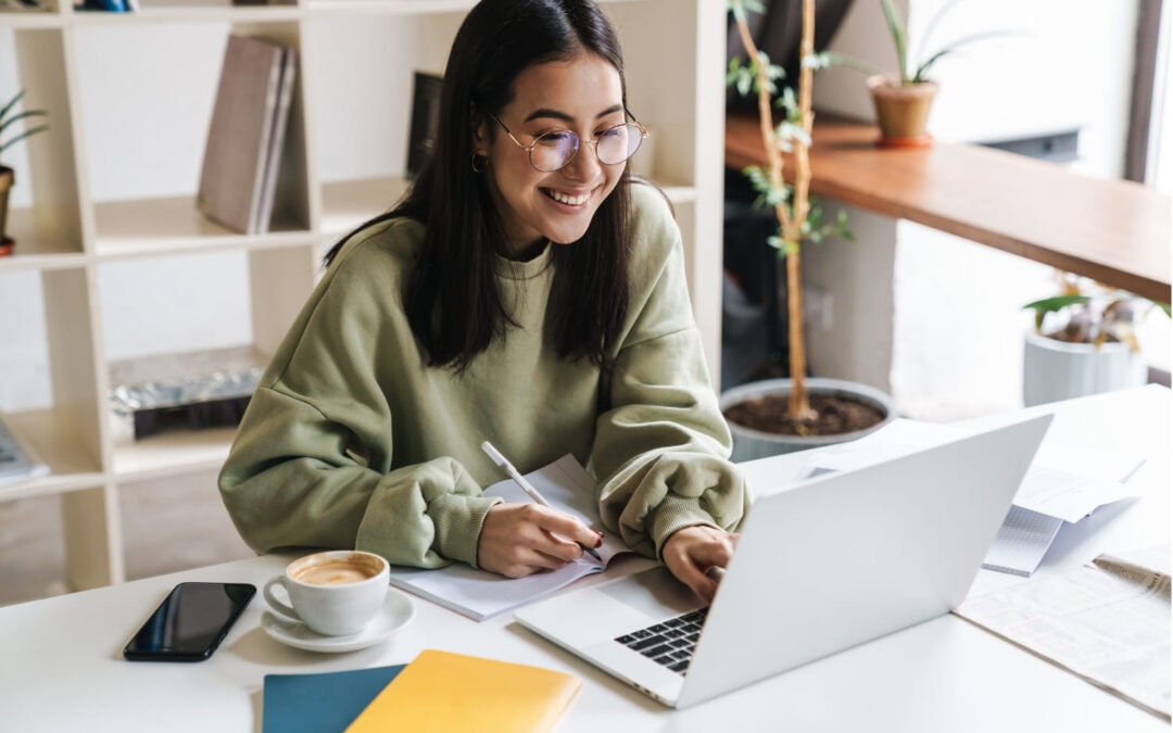 A young woman wearing glasses smiles while studying at a desk with a laptop, notebook, coffee cup, and books. She is writing in a notebook and appears to be working in a bright, modern room with shelves and plants.