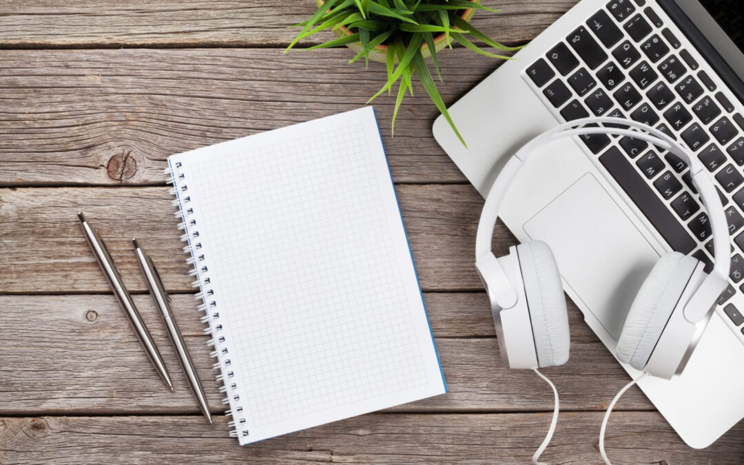 A blank squared notebook, two pens, white headphones, a silver laptop, and a green potted plant on a wooden desk.